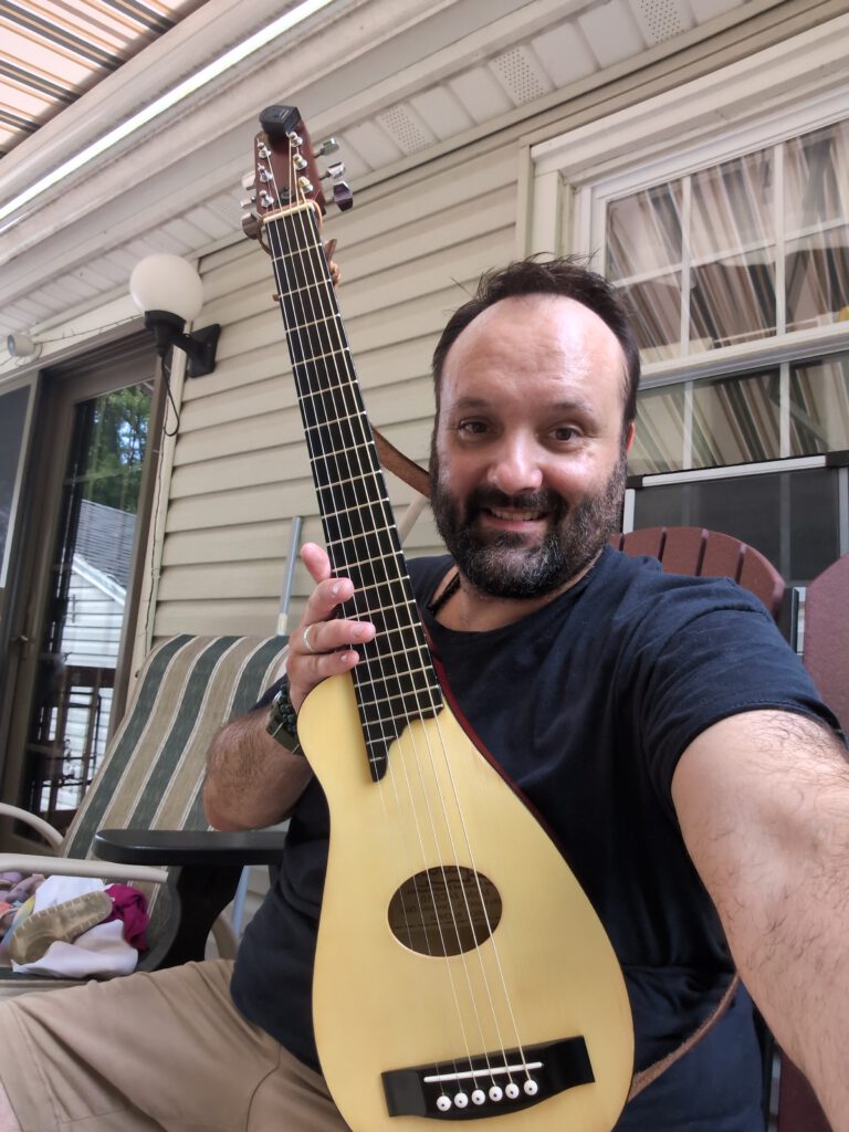 A smiling person sits on a porch holding a small yellow acoustic guitar upright for the camera.