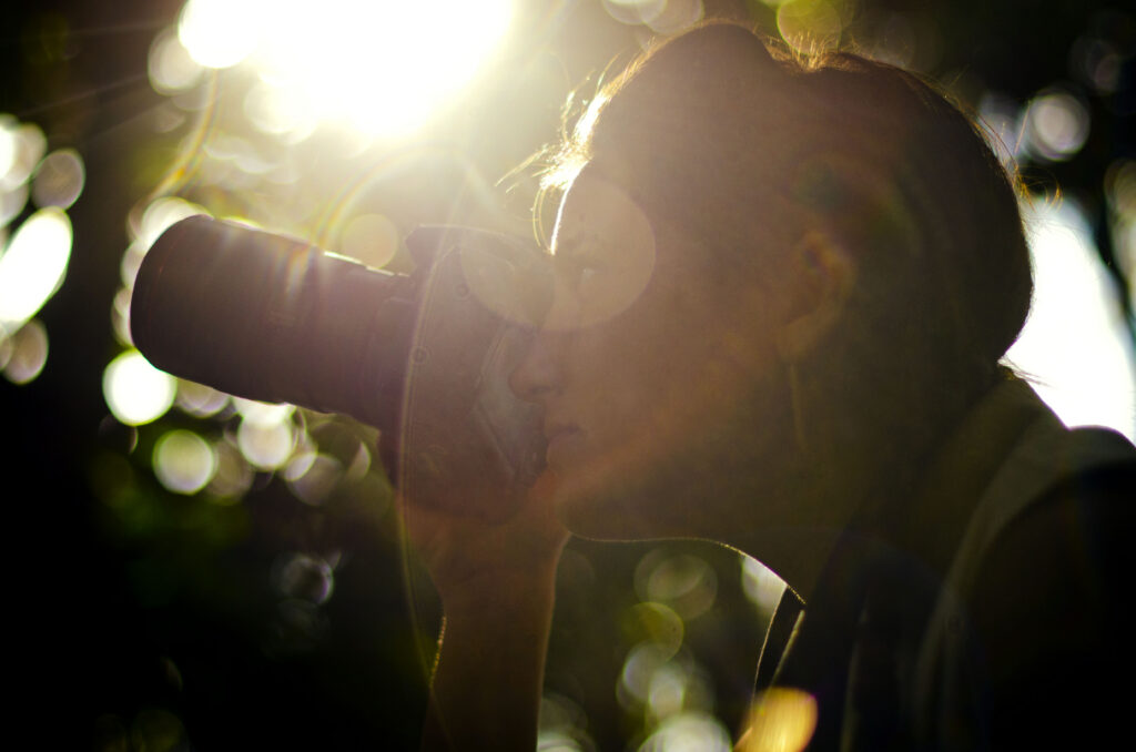 Katie holding a camera up to her eye, framed by warm sunlight and soft bokeh in the background.