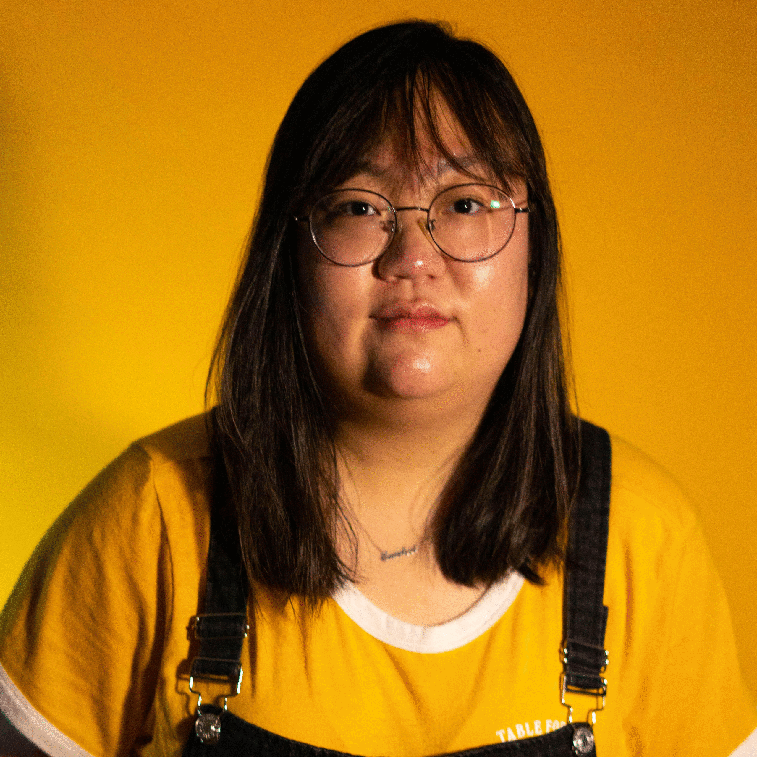A person wearing glasses and black overalls over a yellow shirt stands against a bright yellow background, softly lit for a portrait.