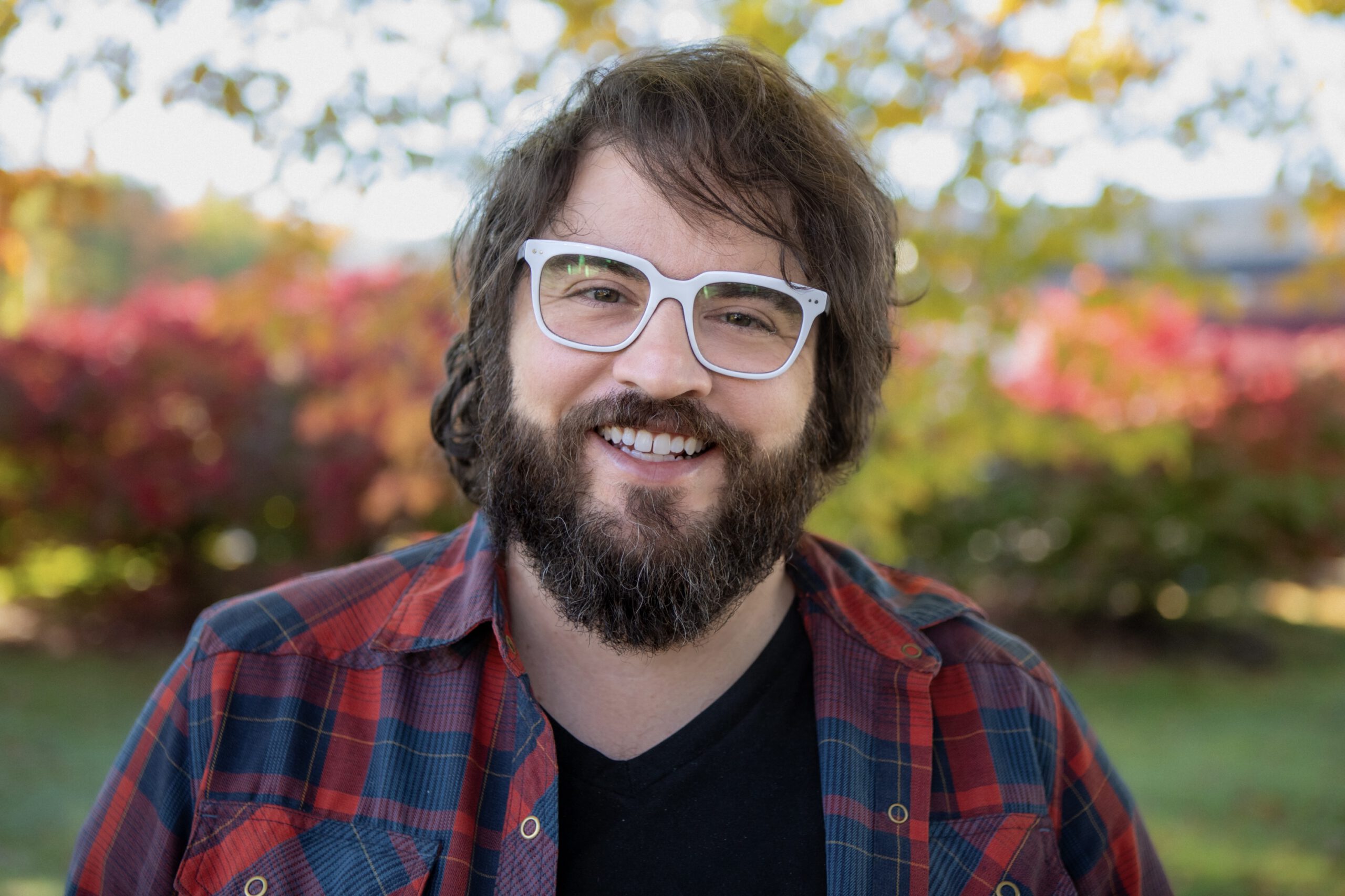 A bearded man in white glasses and a plaid shirt smiles warmly against a colorful background of autumn leaves.