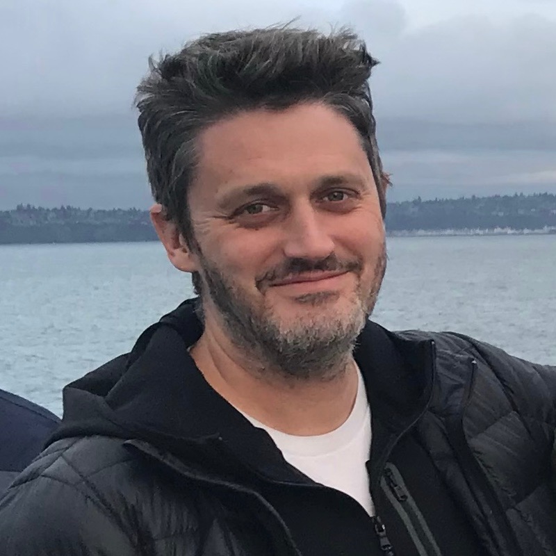 A person standing near a body of water smiles at the camera, with cloudy skies and distant shoreline in the background.