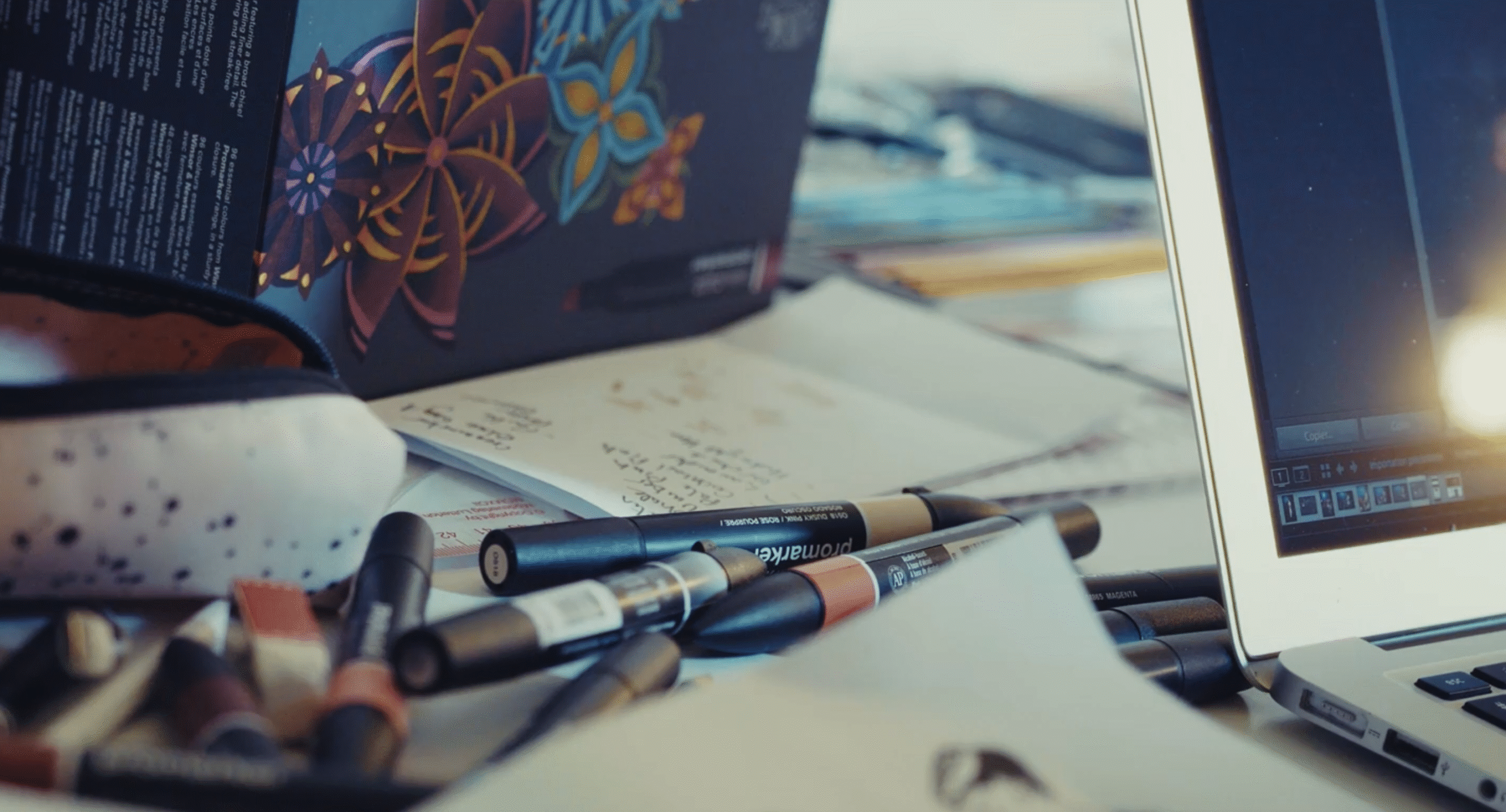 A creative workspace with markers, notes, and a laptop on a desk, softly lit by natural light.